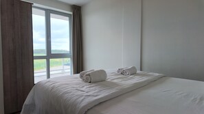 A hotel room with a large bed, two rolled towels, a window with curtains, and a view of a parking area.