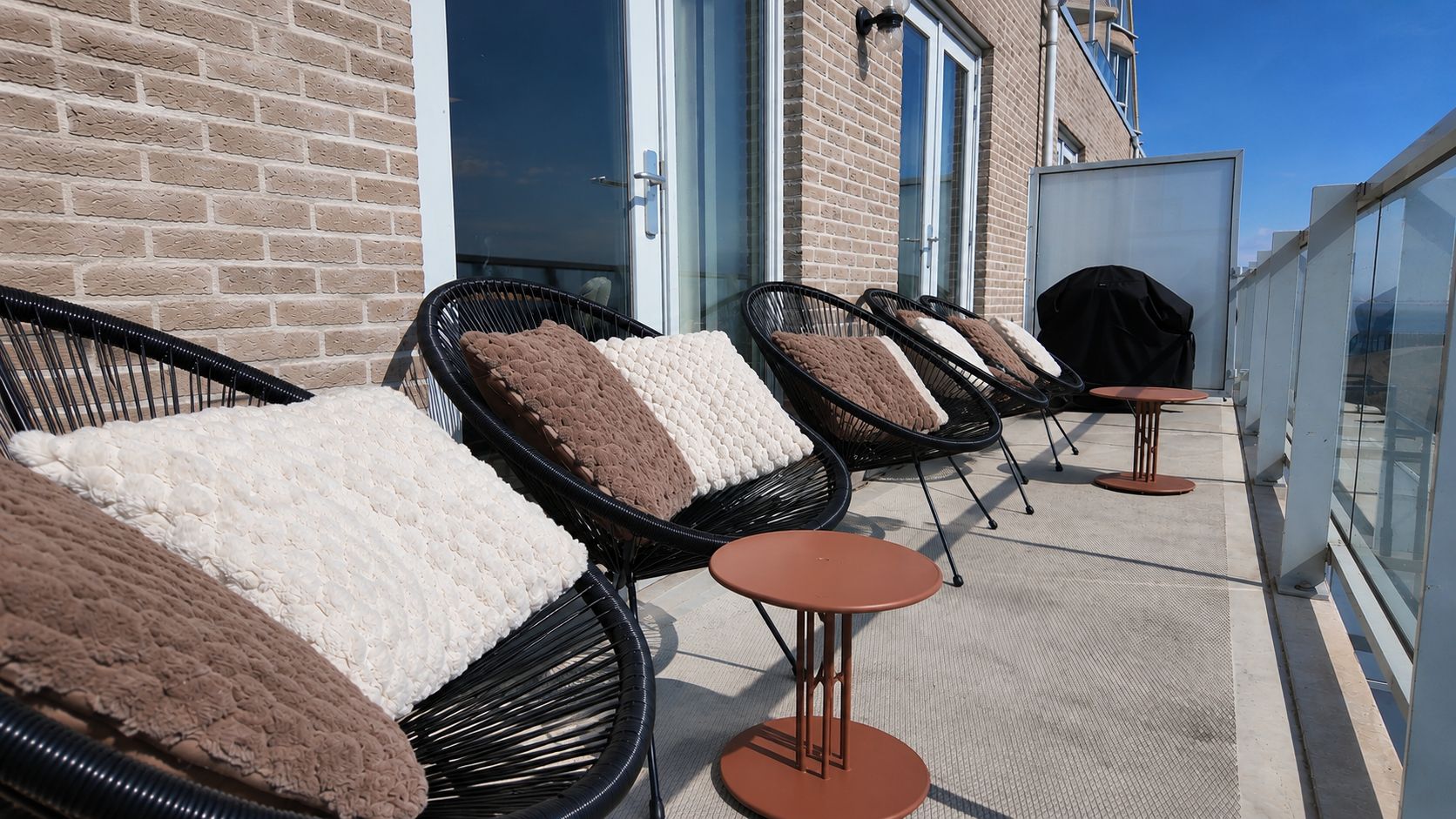 Buiten zitplaatsen met beklede stoelen en kleine ronde tafels, op een balkon met een bakstenen muur en glazen deuren op de achtergrond.