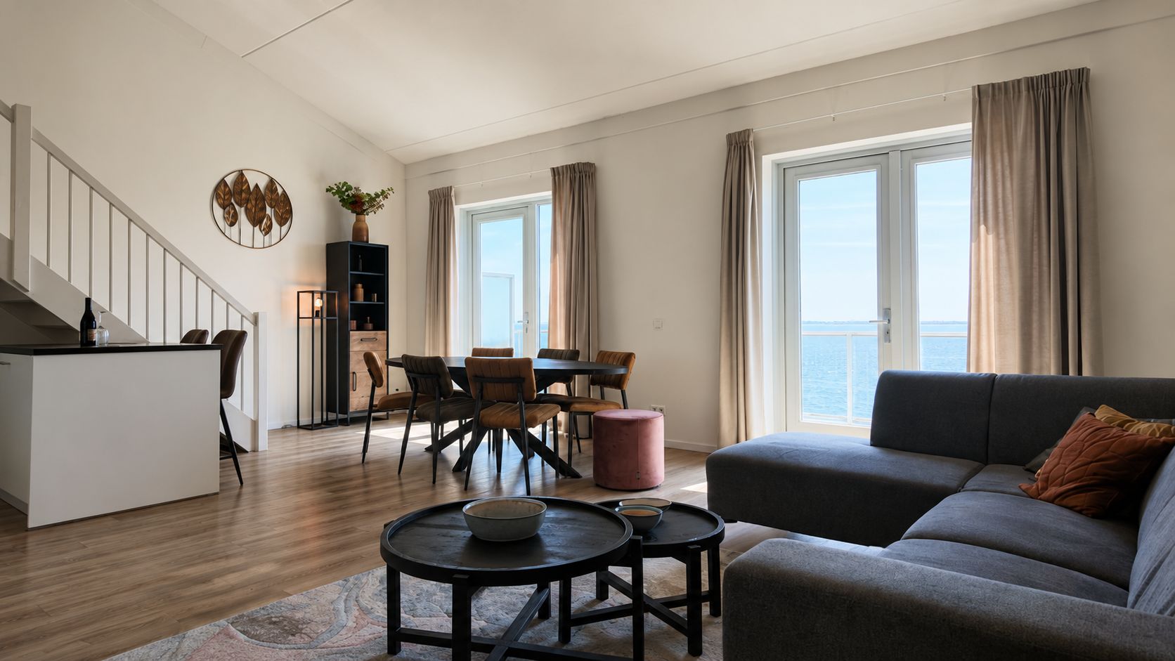 Een moderne woonkamer met een bank, eettafel en stoelen, een keuken met een aanrecht en kasten, en een trap naar een hoger gelegen verdieping.