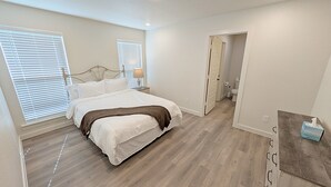 A bedroom with a bed, a nightstand, a lamp, and a door leading to a bathroom. - Lubbock Launchpad Retreat (Lubbock)