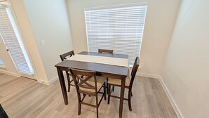 A dining area with a wooden table and chairs, white blinds on the windows, and light-colored walls. - Lubbock Launchpad Retreat (Lubbock)