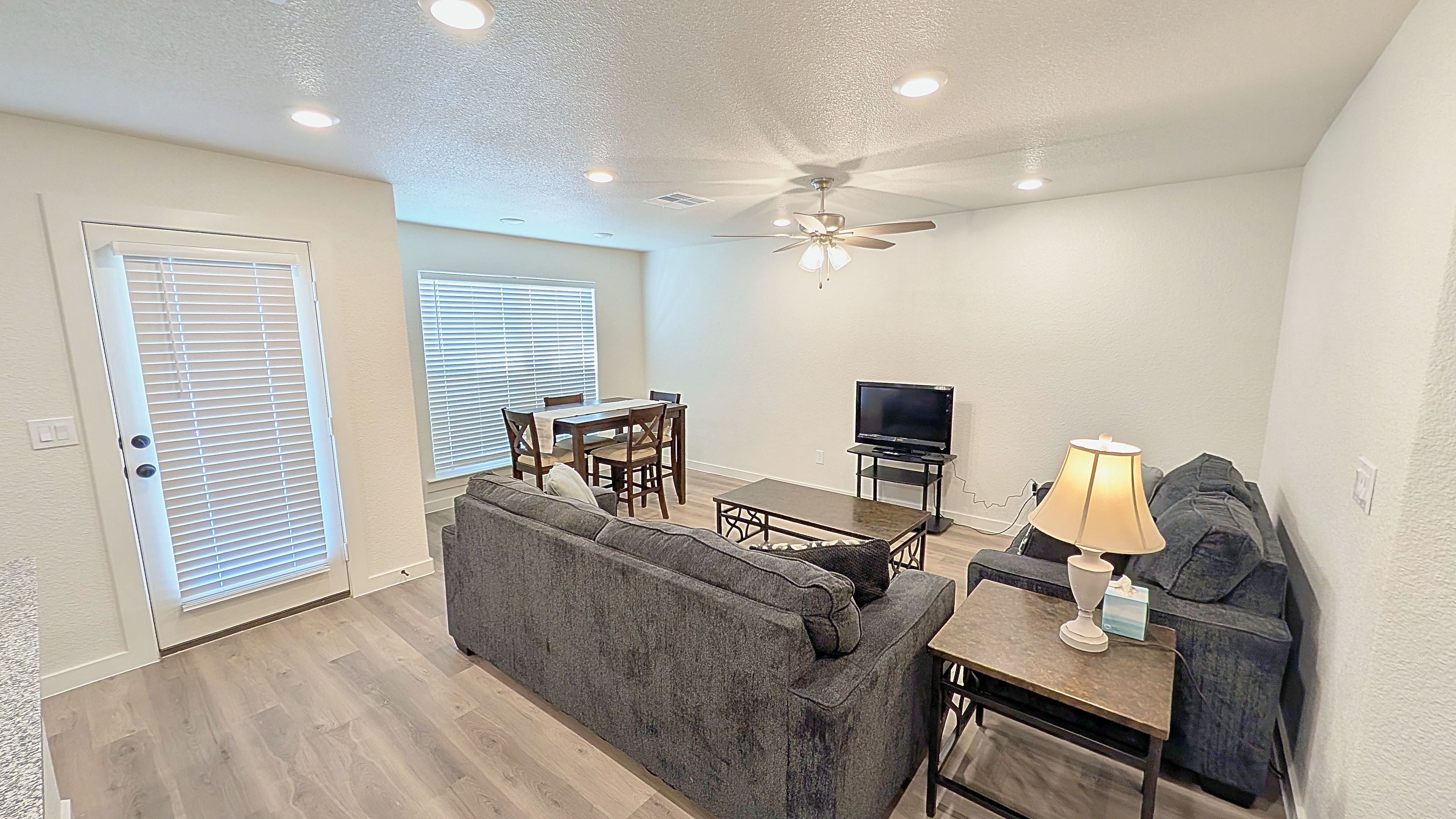 A living room with a grey sofa, a dining table with chairs, a ceiling fan, and a television.