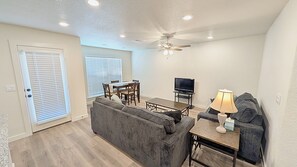 A living room with a grey sofa, a dining table with chairs, a ceiling fan, and a television.
