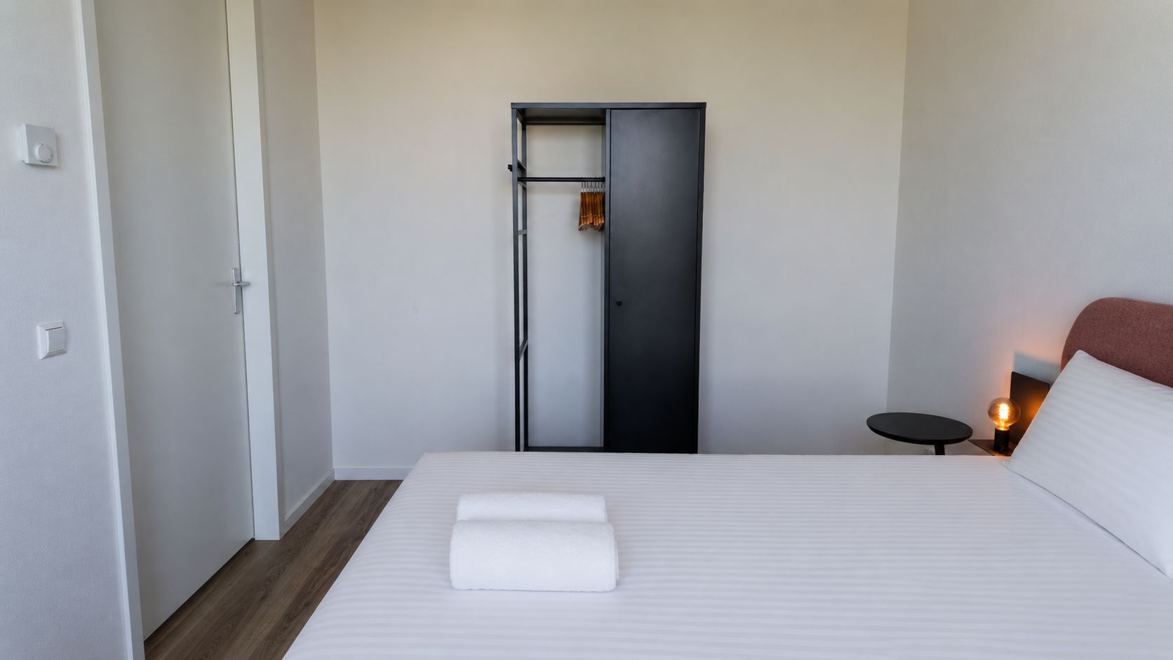 Una camera d'albergo con un letto, un comodino con una lampada, un armadio e una porta.