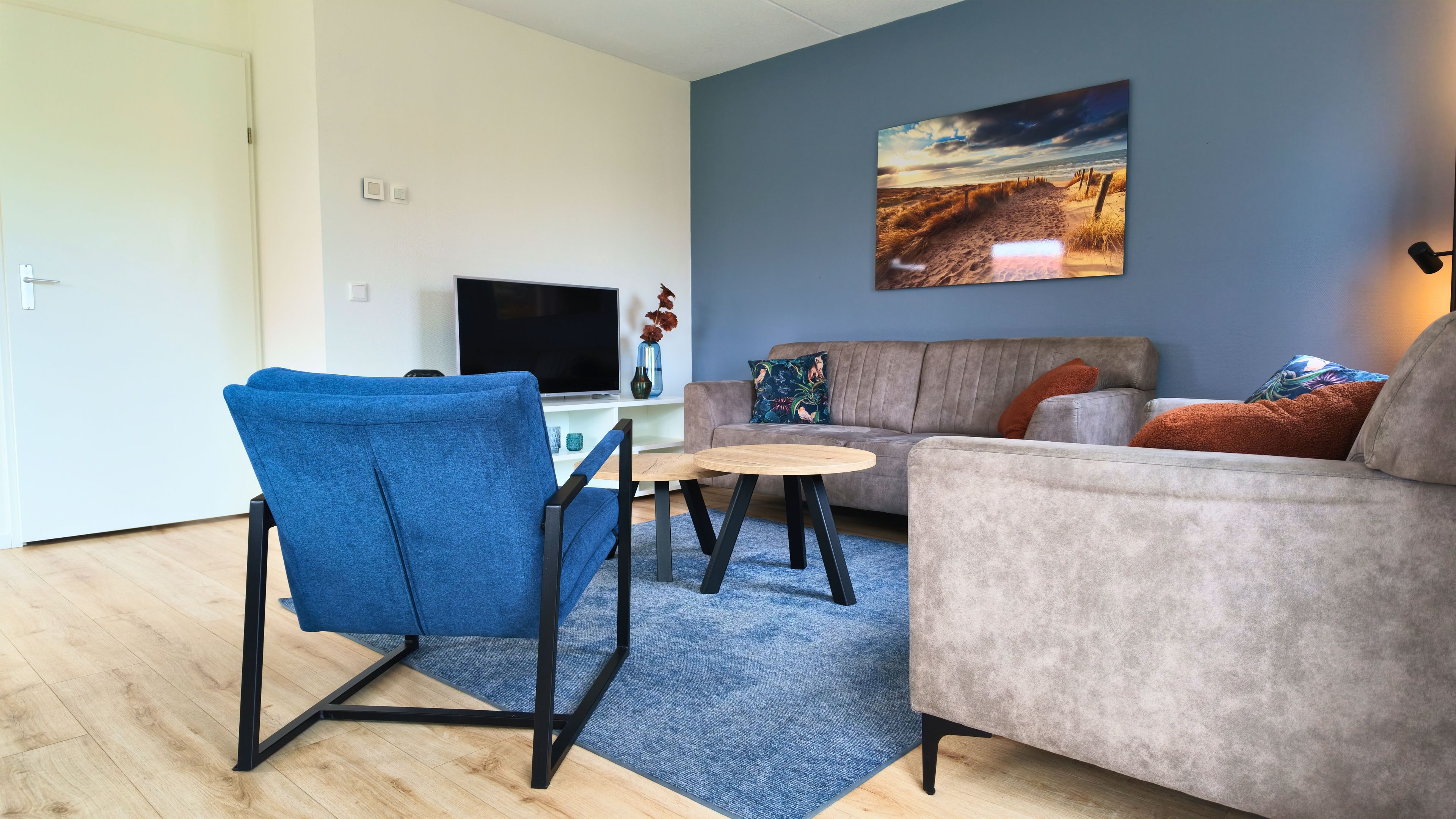 Una sala de estar con un sillón azul, un sofá gris, una mesita redonda y un televisor de pantalla plana.