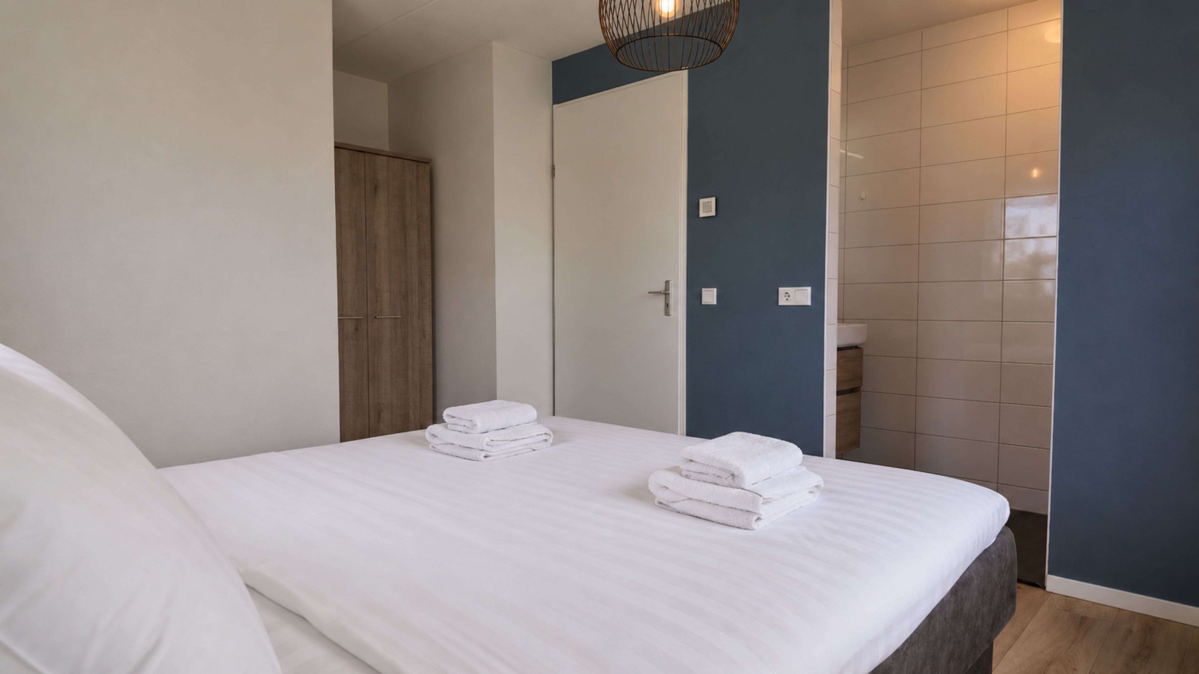 Une chambre d’hôtel avec un lit, une armoire en bois, une salle de bain avec un lavabo et un mur d’accent bleu.