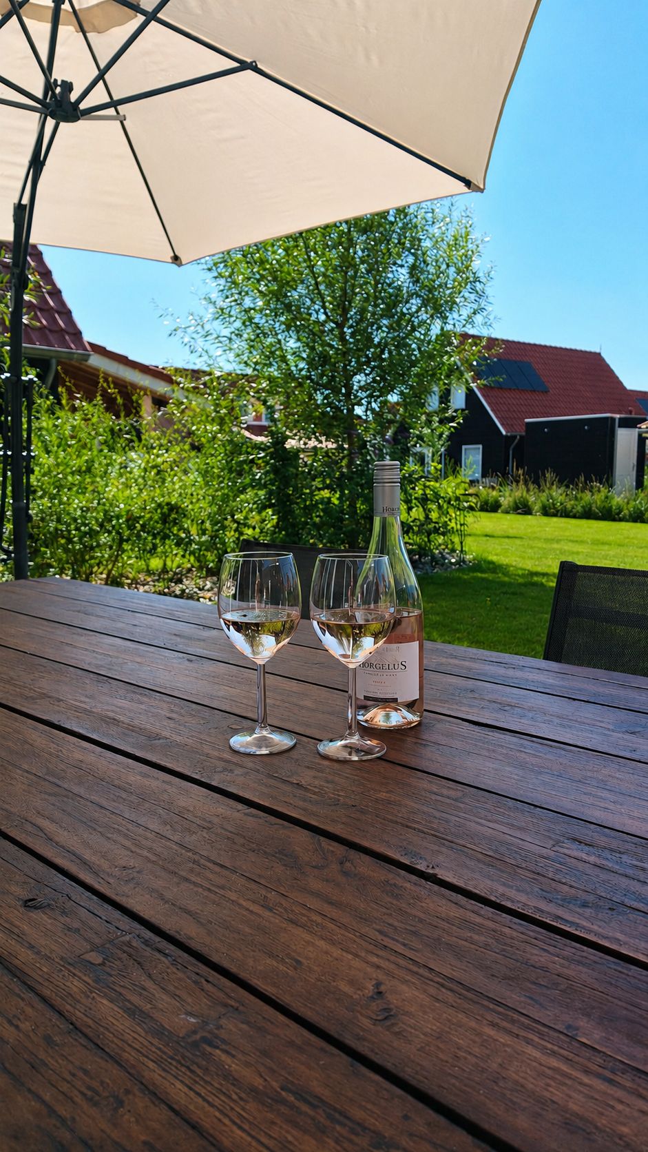 Une table en bois, avec deux verres à vin et une bouteille de vin, placée sous un grand parasol, dans un jardin.