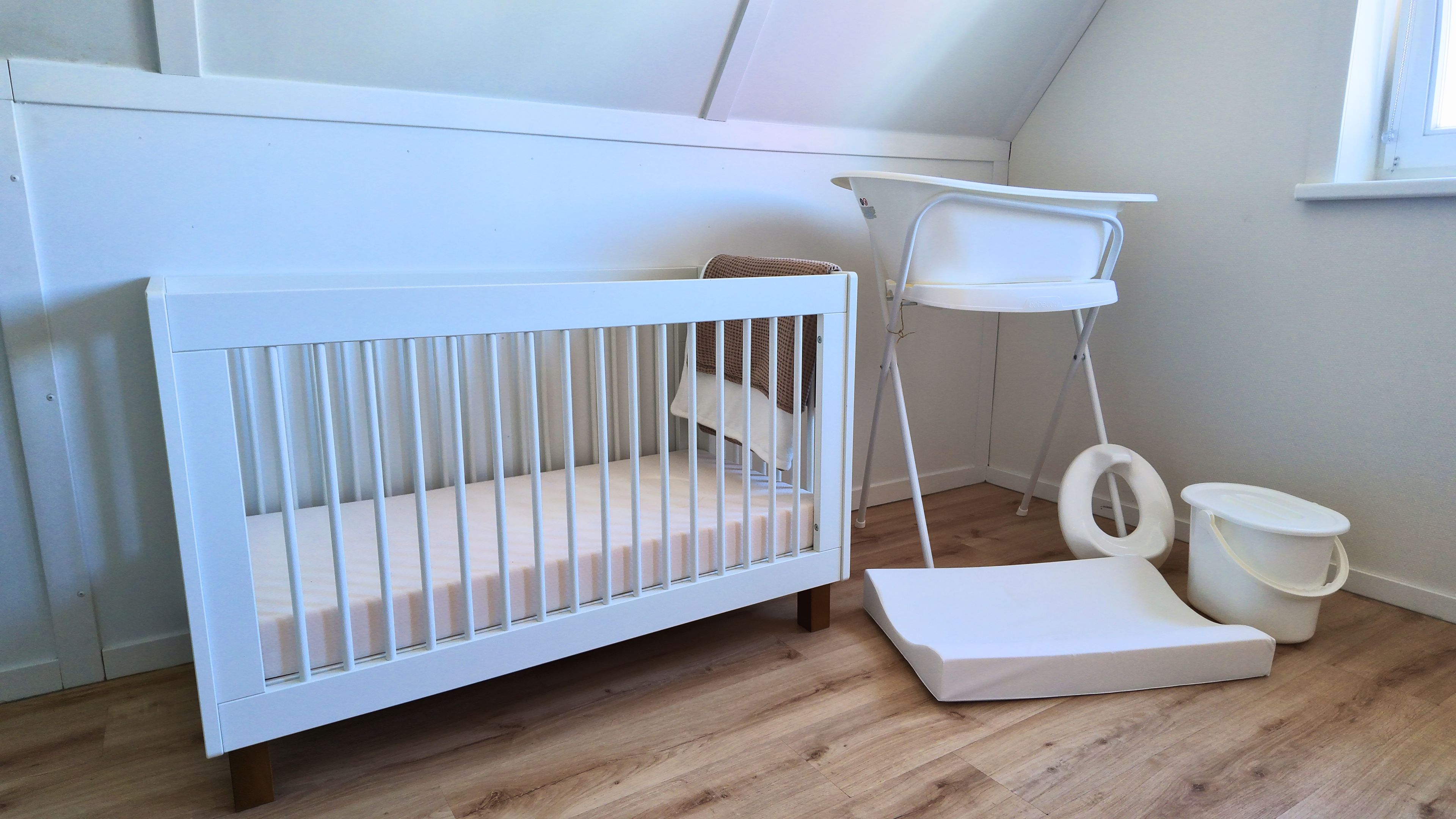 A white crib with a changing table and a storage bin in a room with wooden flooring.