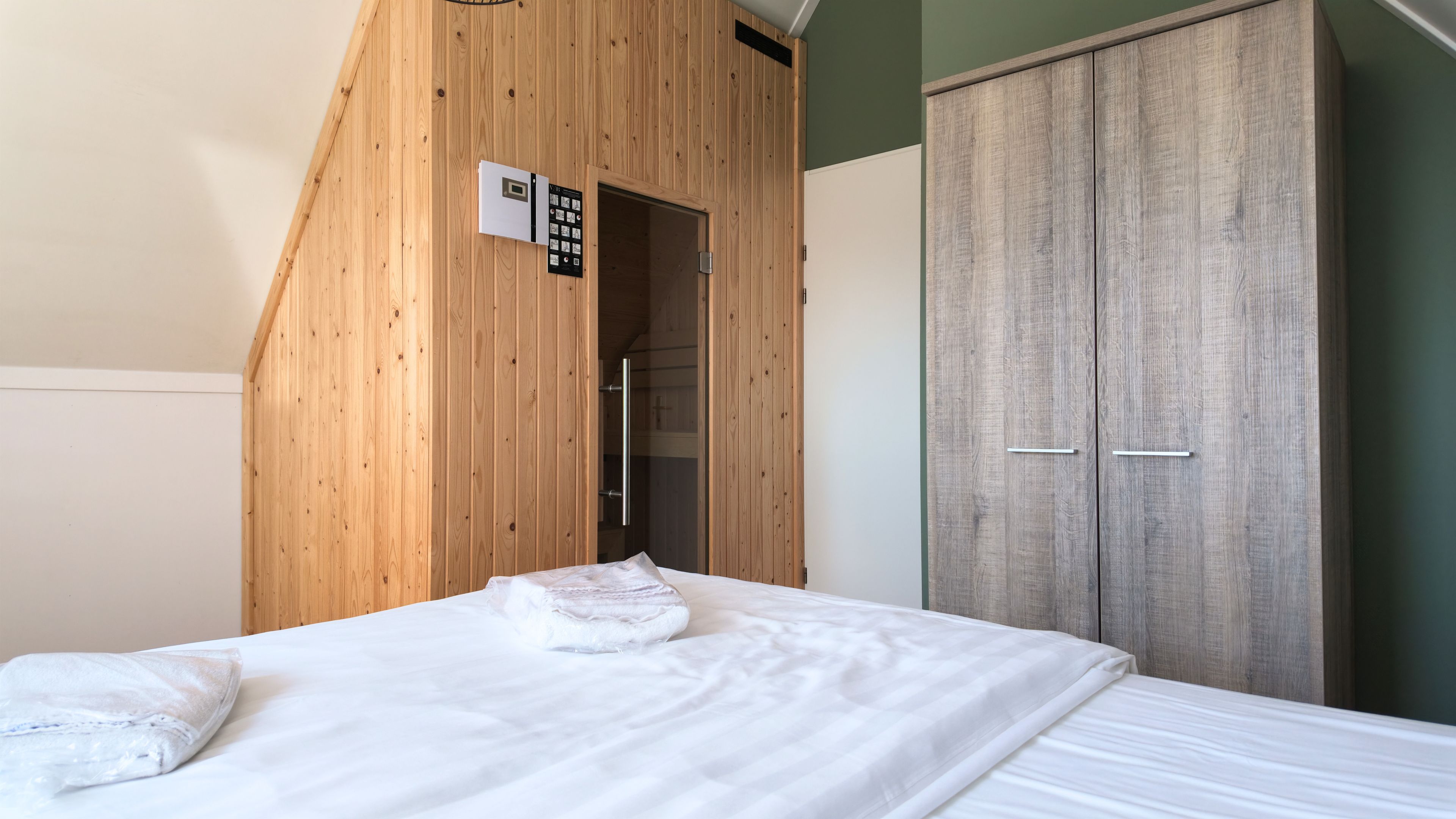 Un dormitorio con techo inclinado de madera, una cama con ropa blanca y un armario de dos puertas.