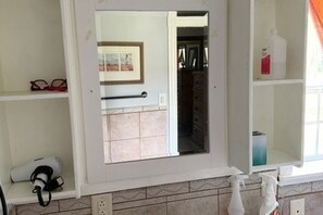Un baño con espejo, toallero, estante con una botella y ventana con cortinas.