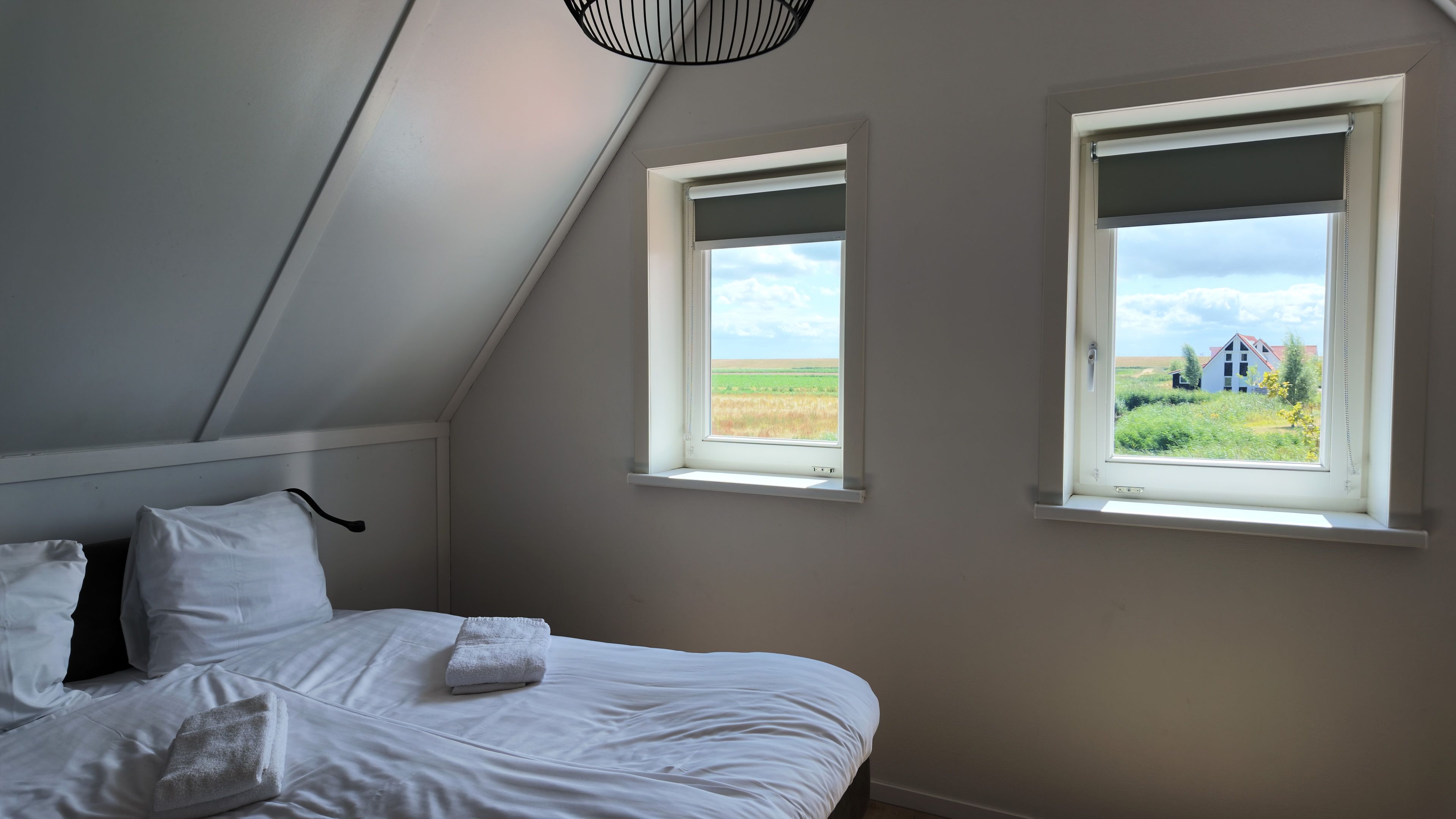 Una camera da letto con soffitto spiovente, due finestre che si affacciano su un campo e un edificio, un letto con lenzuola bianche e un lampadario.