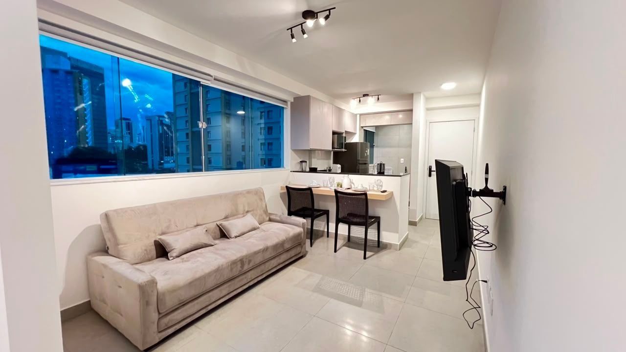 Un salon moderne avec un canapé, un coin repas avec des chaises, une cuisine équipée d’armoires et une grande fenêtre donnant sur la ville.