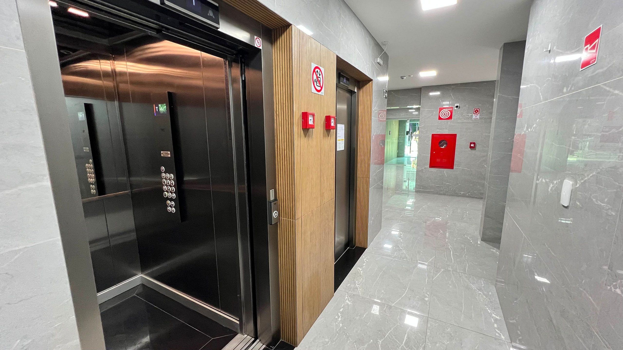 Un ascensor moderno con detalles metálicos y paneles de madera, acompañado por botones de emergencia en rojo y una indicación clara del piso.