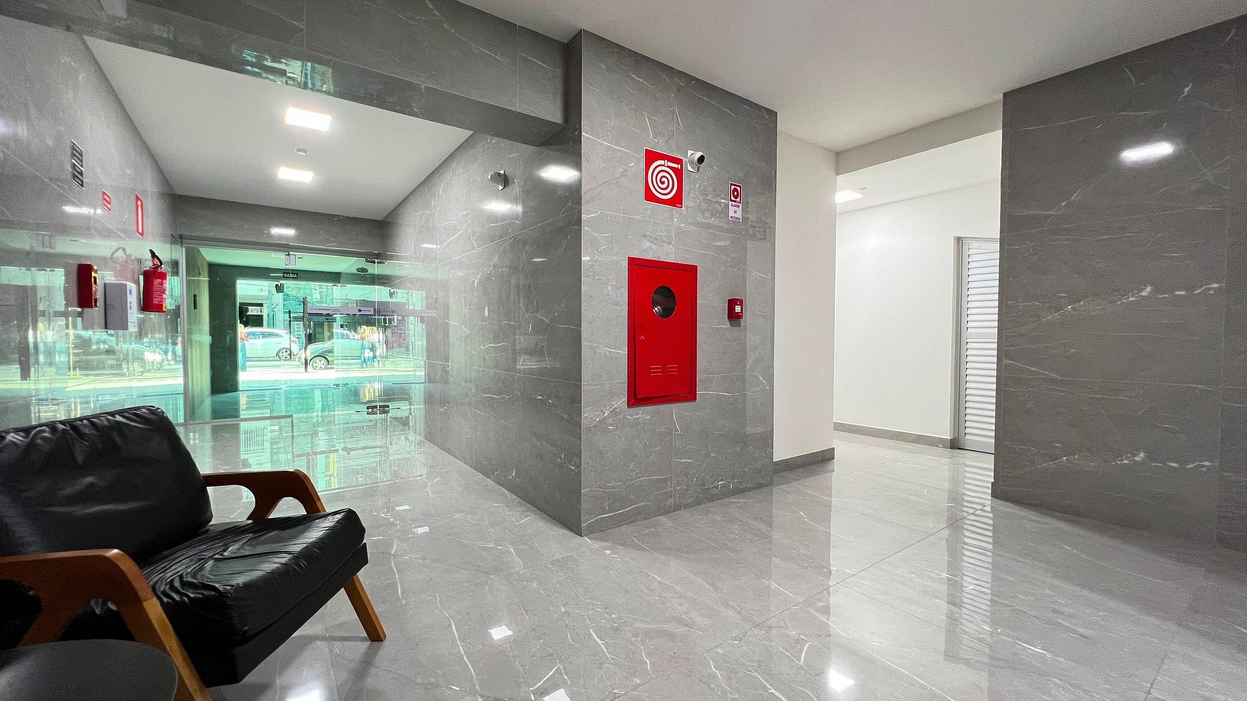 Un vestíbulo moderno con suelo de mármol, un letrero de salida de emergencia en rojo y un escaparate de vidrio al fondo.