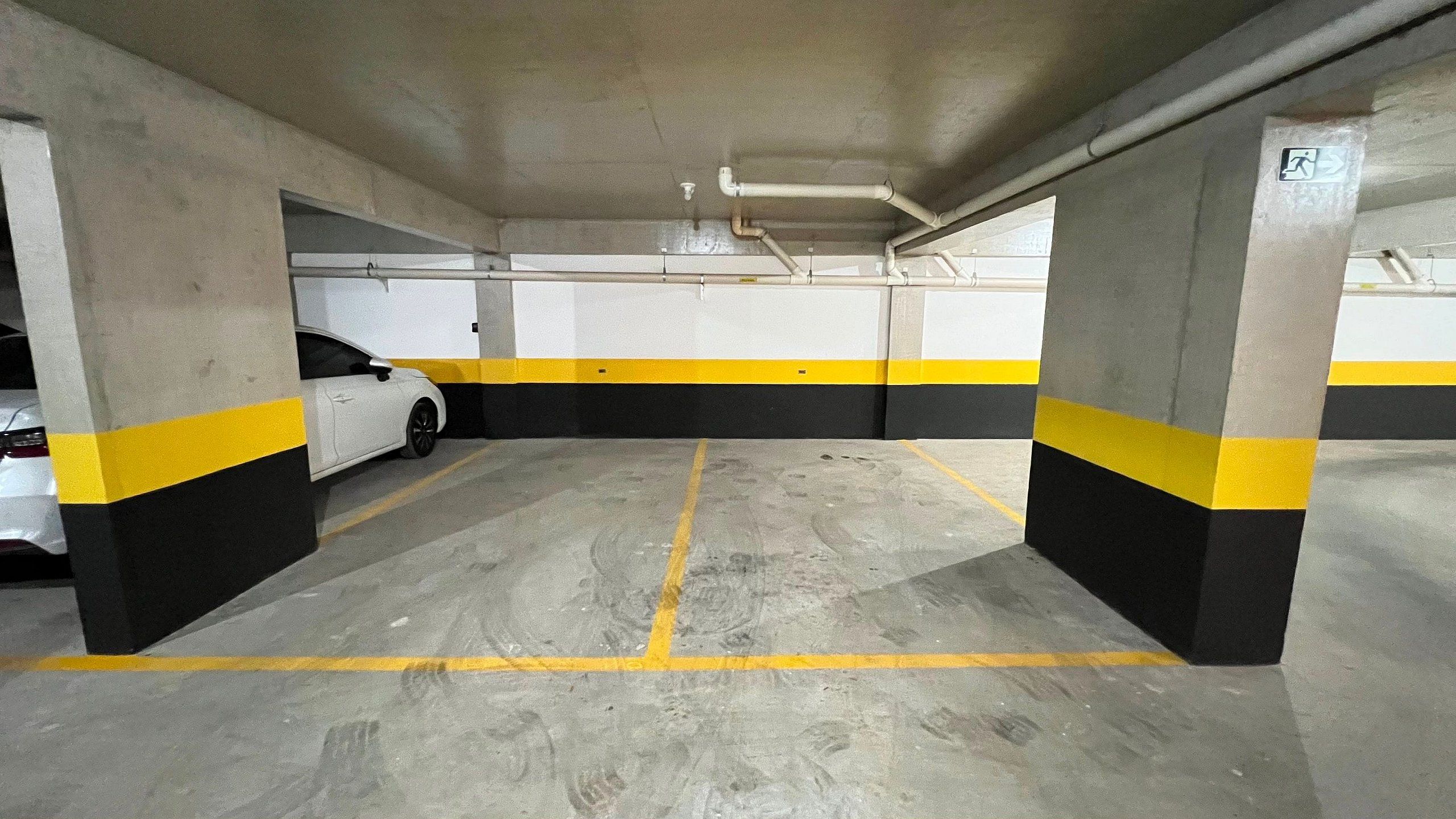 Un parking avec des piliers en béton, des lignes peintes en jaune et en noir, et une voiture blanche garée dans une place.