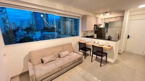 Un salon moderne comprenant un canapé, une table à manger avec des chaises, un coin cuisine et une grande fenêtre donnant sur la ville.