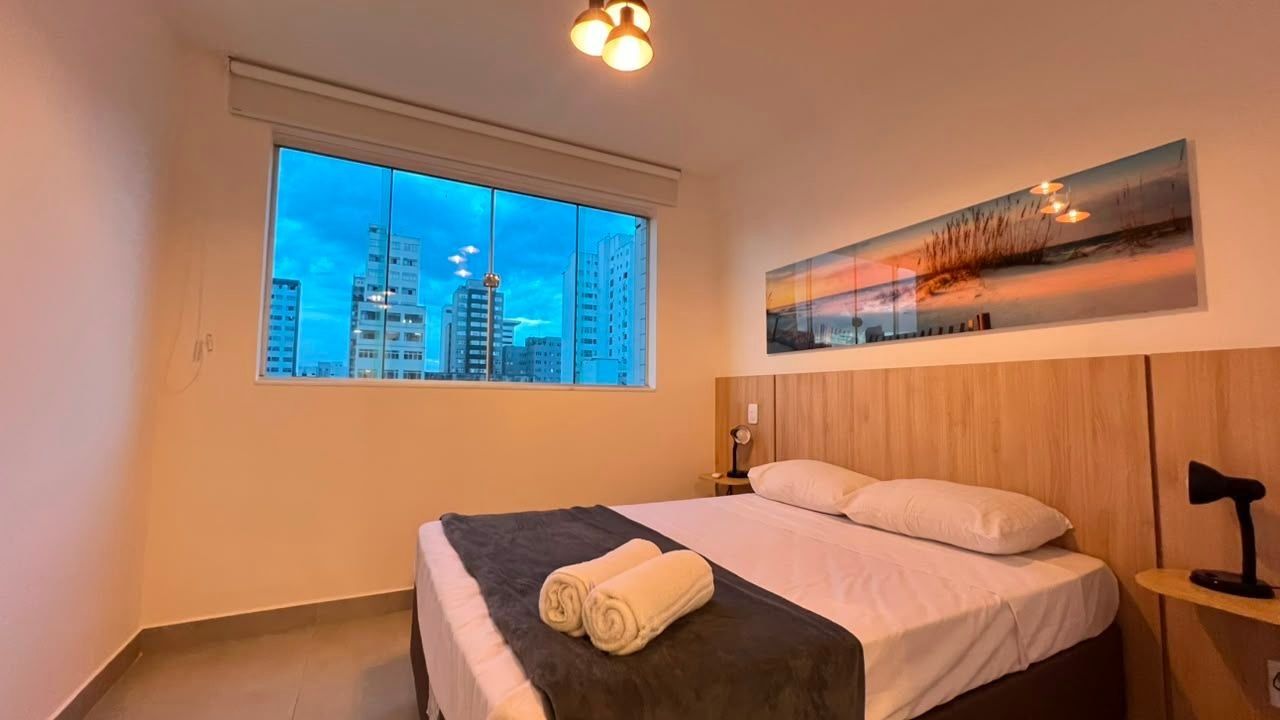 Une chambre d’hôtel avec une grande fenêtre, un lit recouvert de draps blancs, un plaid noir, un tableau accroché au mur et une table de chevet avec une lampe.