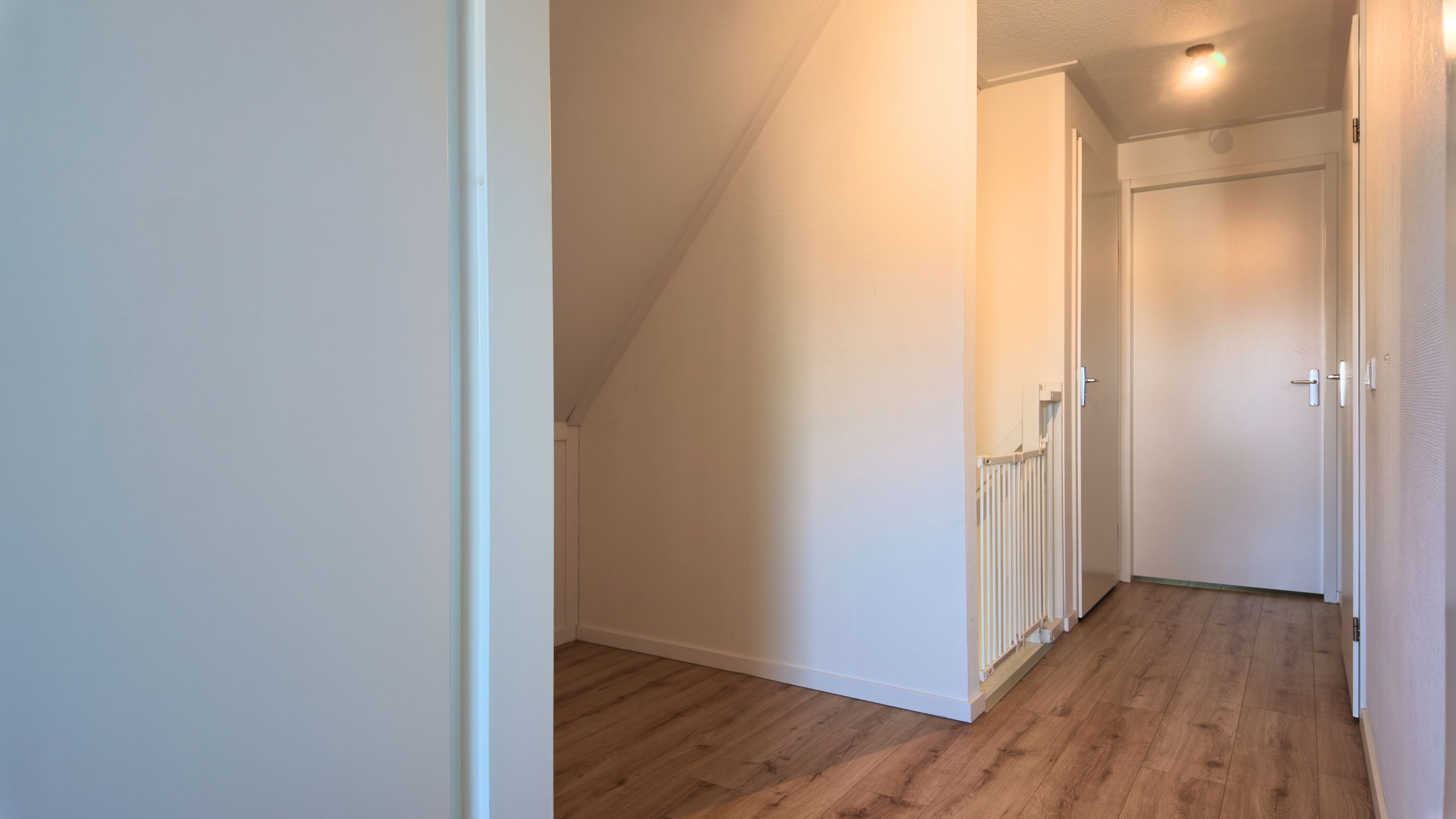 Un couloir étroit et bien éclairé, doté d’un plancher en bois et de murs blancs, qui mène à une porte au bout.