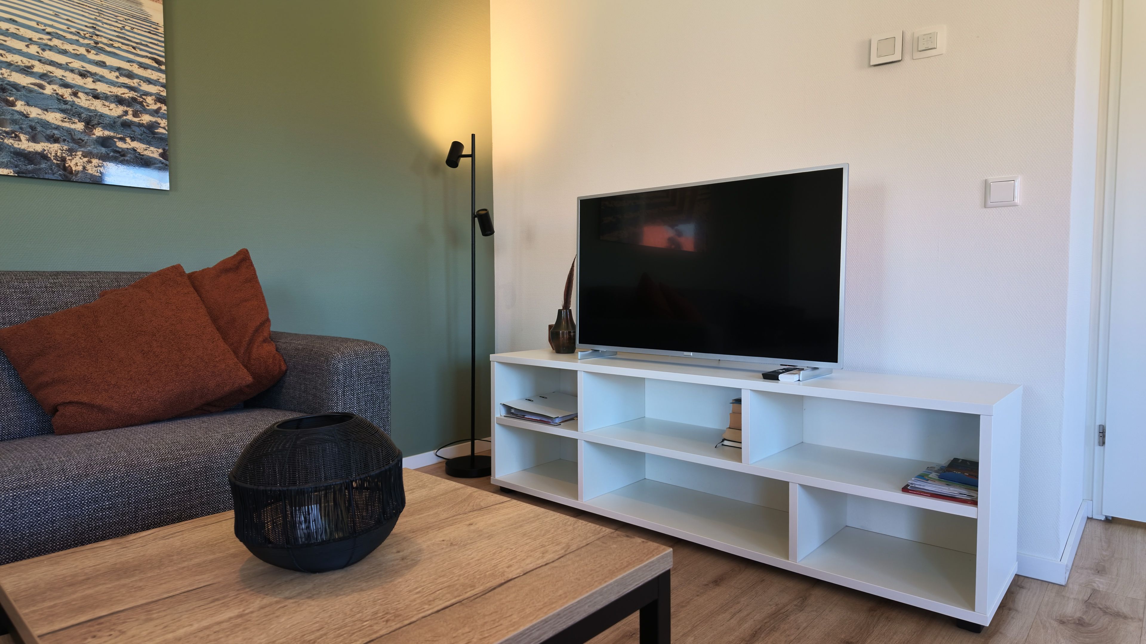 Un salon avec un canapé, une table basse, un téléviseur posé sur un meuble blanc et un lampadaire.