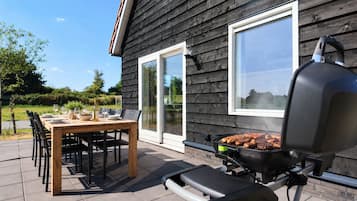 Une terrasse extérieure avec une table en bois dressée pour les repas, un barbecue et une maison aux bardages sombres et aux grandes fenêtres.