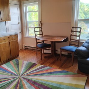 A room with a wooden table and two chairs, a colorful rug, a wooden cabinet, and a window with a view of trees. - Superb 3-bedroom apartment   in quaint Jay Maine (Jay)