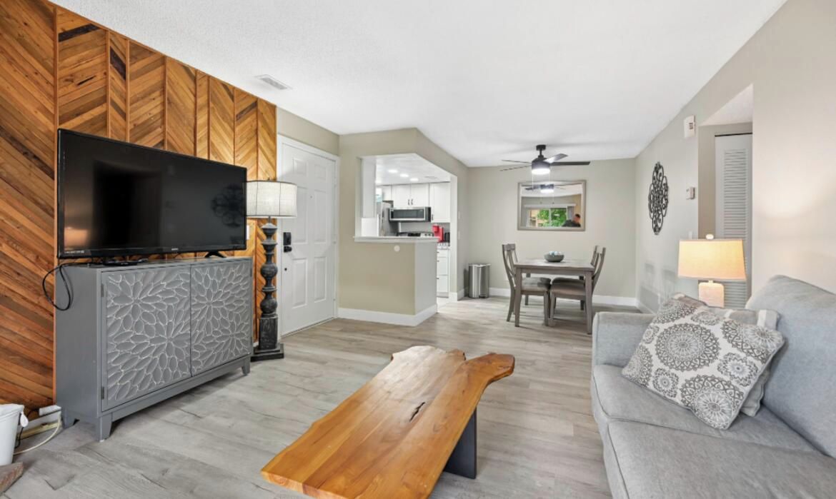 A living room with a flat-screen TV, grey patterned cabinet, wooden coffee table, grey sofa with patterned cushions, and a dining area with a table and chairs.