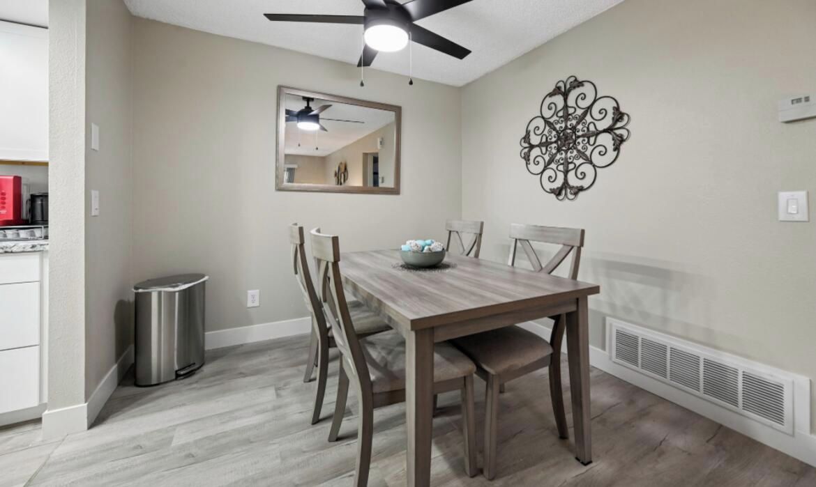 A dining area with a wooden table, chairs, a ceiling fan, a wall-mounted mirror, and a decorative wall piece.