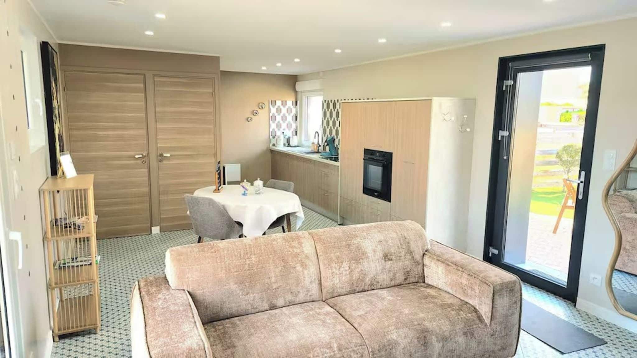 Een woonkamer met een bank, een eettafel met stoelen, een keukenhoek met een televisie en een glazen schuindeur naar buiten.