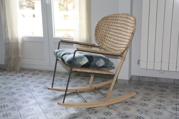A rocking chair with a woven backrest and a cushion, positioned in a room with patterned floor tiles and a white radiator.