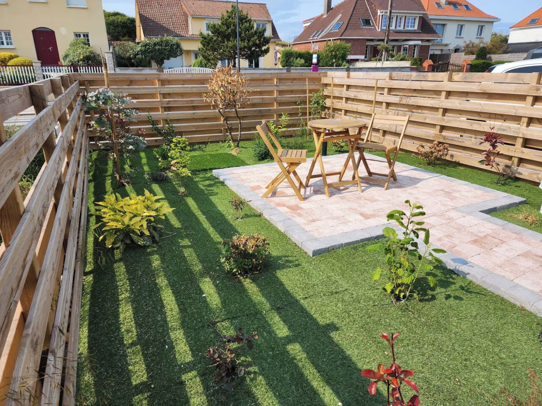 Ein gepflegter Garten mit Holzzaun, einer Terrassenfläche mit Tisch und Stühlen sowie verschiedenen Pflanzen.