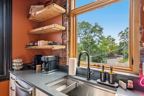 A kitchen with a built-in sink, a coffee machine, and a window with a view of trees and a road. - Dedicated Parking | King Bed | Arcade | Patio (Pittsburgh)