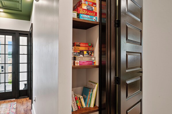 A bookshelf with various board games and puzzles, a door with a mirror, and a view of a cityscape through a window. - Dedicated Parking | King Bed | Arcade | Patio (Pittsburgh)