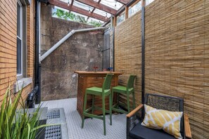 A small outdoor seating area with a wooden table and two chairs, a bench with a cushion, and a bamboo wall. - Dedicated Parking | King Bed | Arcade | Patio (Pittsburgh)