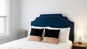 En pænt redt seng med hvide og beige puder, et blåt sengegavl og et lille natbord med en lampe.