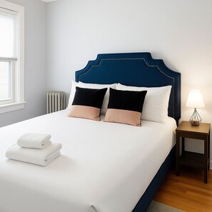 Een net opgemaakt bed met witte en beige kussens, een blauw hoofdbord en een klein nachtkastje met een lamp.