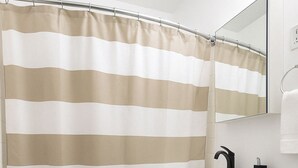 Et badeværelse med cremefarvet bruseforhæng, en hvid vask med sort armatur, et toilet og et spejl over vasken.