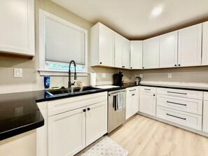 A kitchen with white cabinets, a black countertop, and a window with blinds.