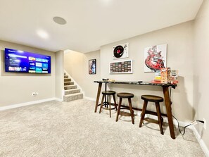 A living room with a flat-screen TV, a staircase, a bar area with stools, and wall decorations including a record and a guitar.