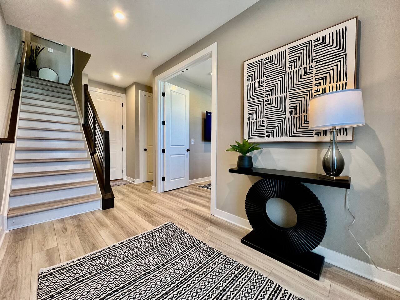 A modern interior with a staircase, a patterned rug, a wall-mounted shelf with a lamp, and a geometric wall art piece.
