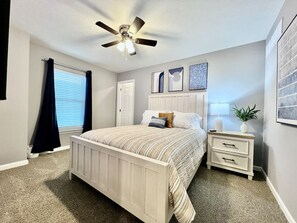 Un dormitorio con cama, mesita de noche, ventilador de techo y ventana con cortinas.
