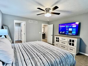 A bedroom with a bed, a ceiling fan, a TV mounted on the wall, and a dresser.