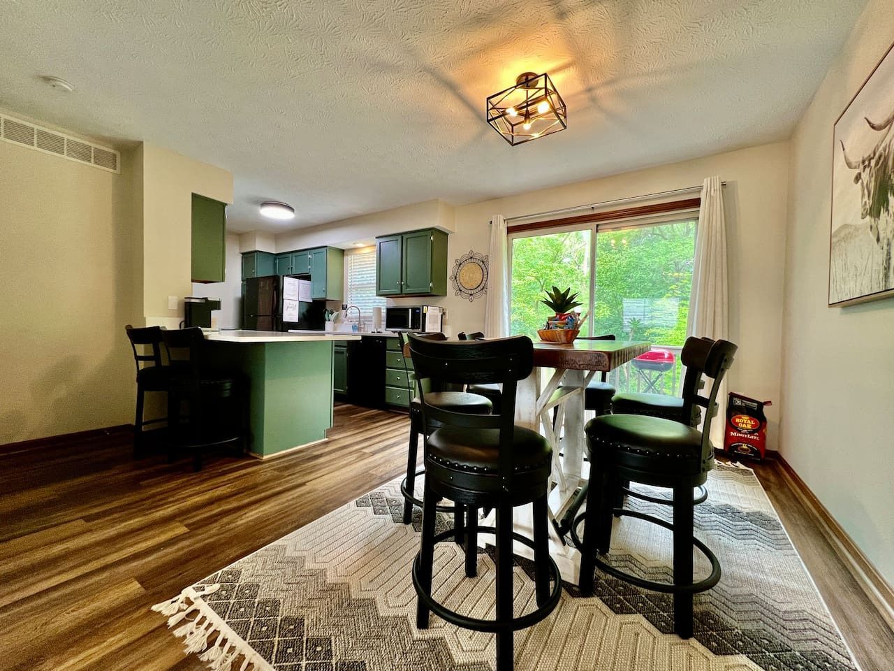A modern kitchen with green cabinets, a dining area with black chairs, and a large window with a view of greenery.