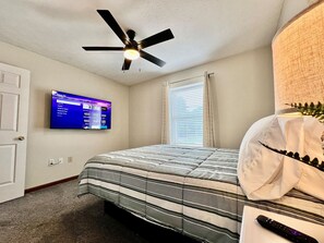 A bedroom with a bed, a ceiling fan, a TV mounted on the wall, and a window with curtains.