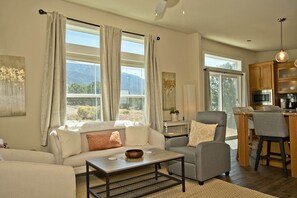 A spacious living room with a sofa, armchair, and coffee table. A kitchen area with wooden cabinets and a bar stool is visible in the background. - Two Suite Mountain Retreat (Saguache County)