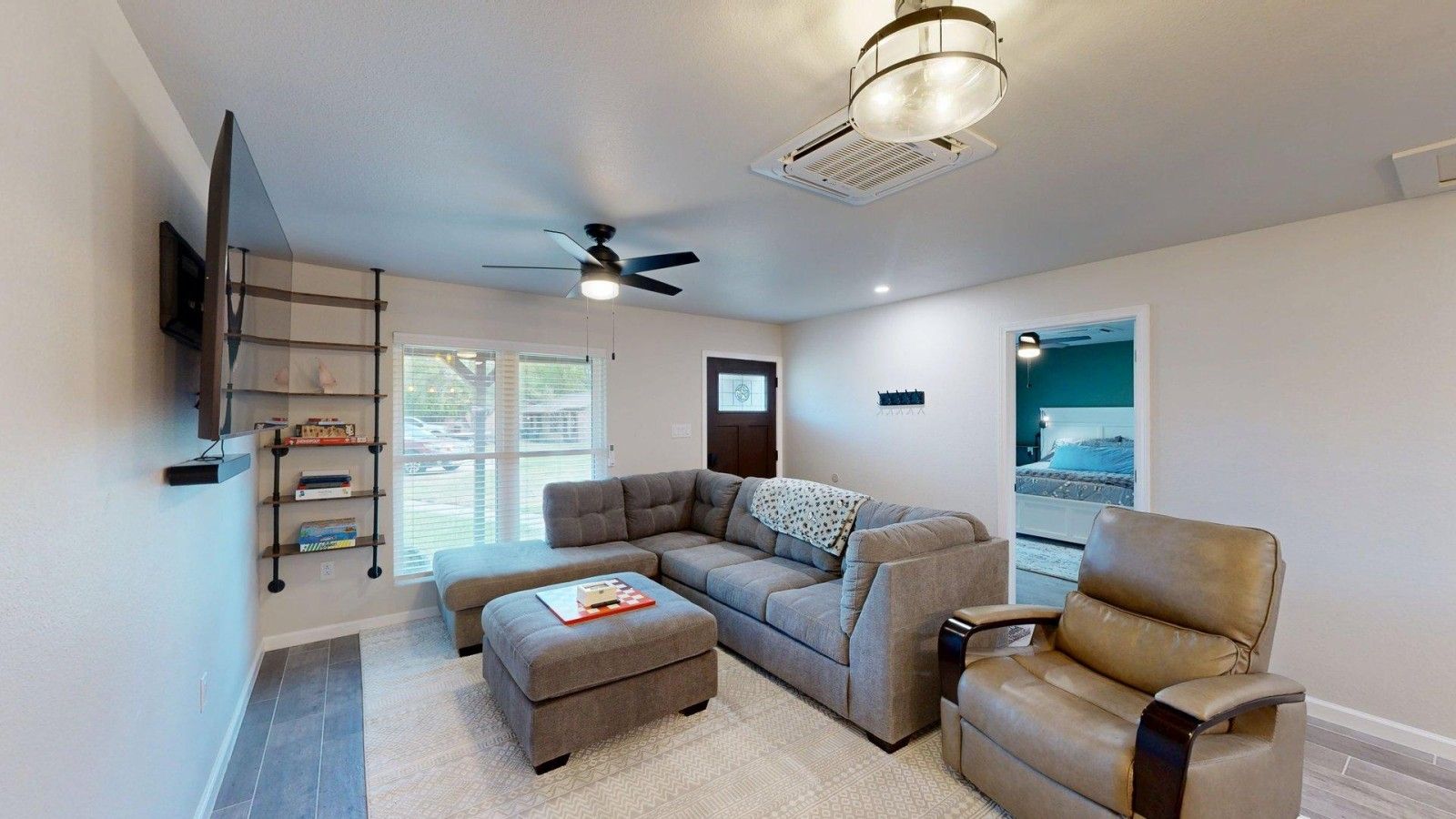 A modern living room with a sectional sofa, a leather armchair, a flat-screen TV mounted on the wall, and a ceiling fan.