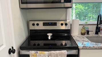 A kitchen with a stainless steel Whirlpool microwave, a black stove with a digital clock, and a window with a view of trees.