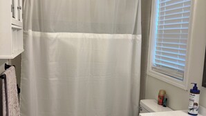 Un baño con cortina blanca de ducha, inodoro, lavamanos con artículos de aseo y ventana con persianas.