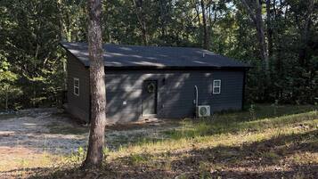 A small, single-story building with a dark roof and a door, surrounded by trees and a grassy area.