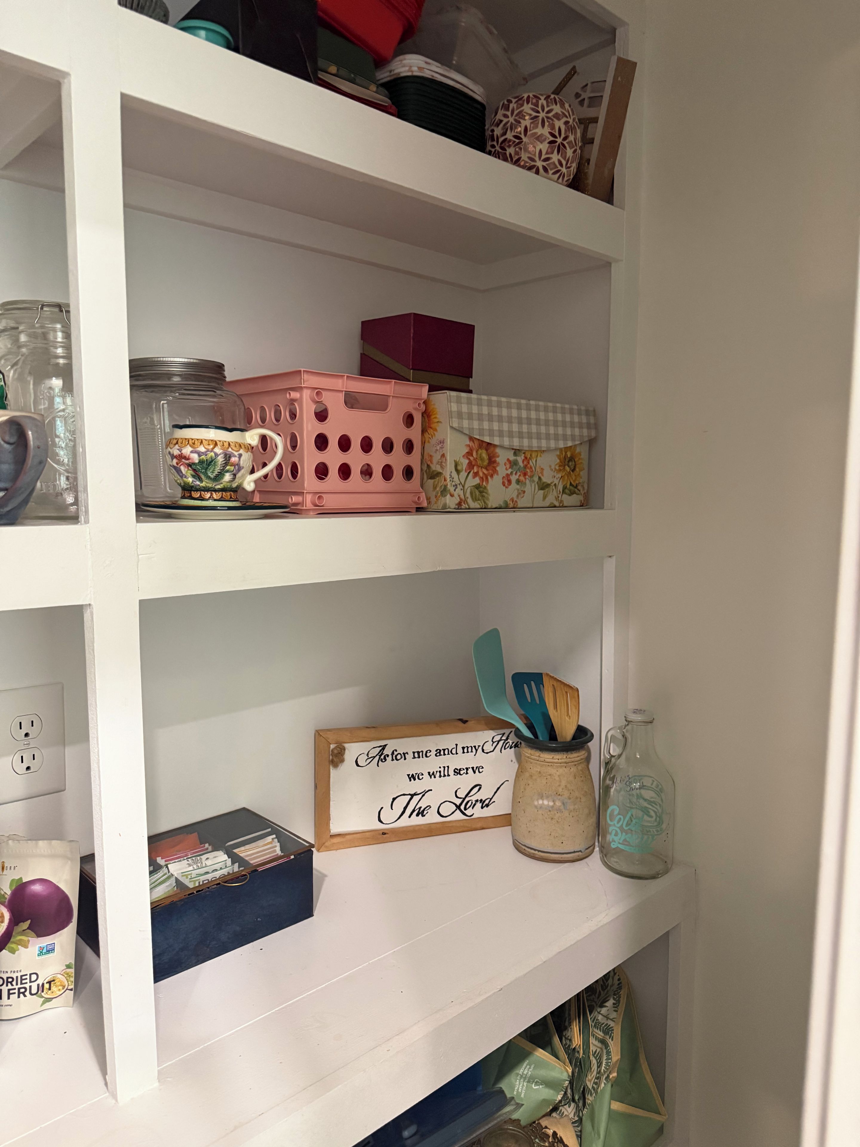 Valkoinen hylly, jossa on erilaisia keittiötarvikkeita, kuten vaaleanpunainen kori, kukkakuviollinen rasia ja puinen kyltti, jossa on uskonnollinen viesti.