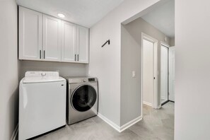 A laundry room with a washing machine and dryer, white cabinets, and a hallway leading to another room. - Family Haven | Theater | Game Room | Backyard Fun (Lawrenceville)